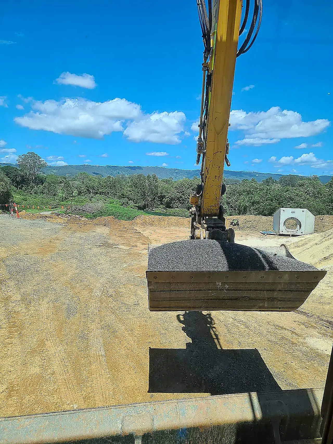 Yellow excavator carrying a bucket of crushed aggregate with a precast concrete pit on an SEQ civil site, photographed from the cab.