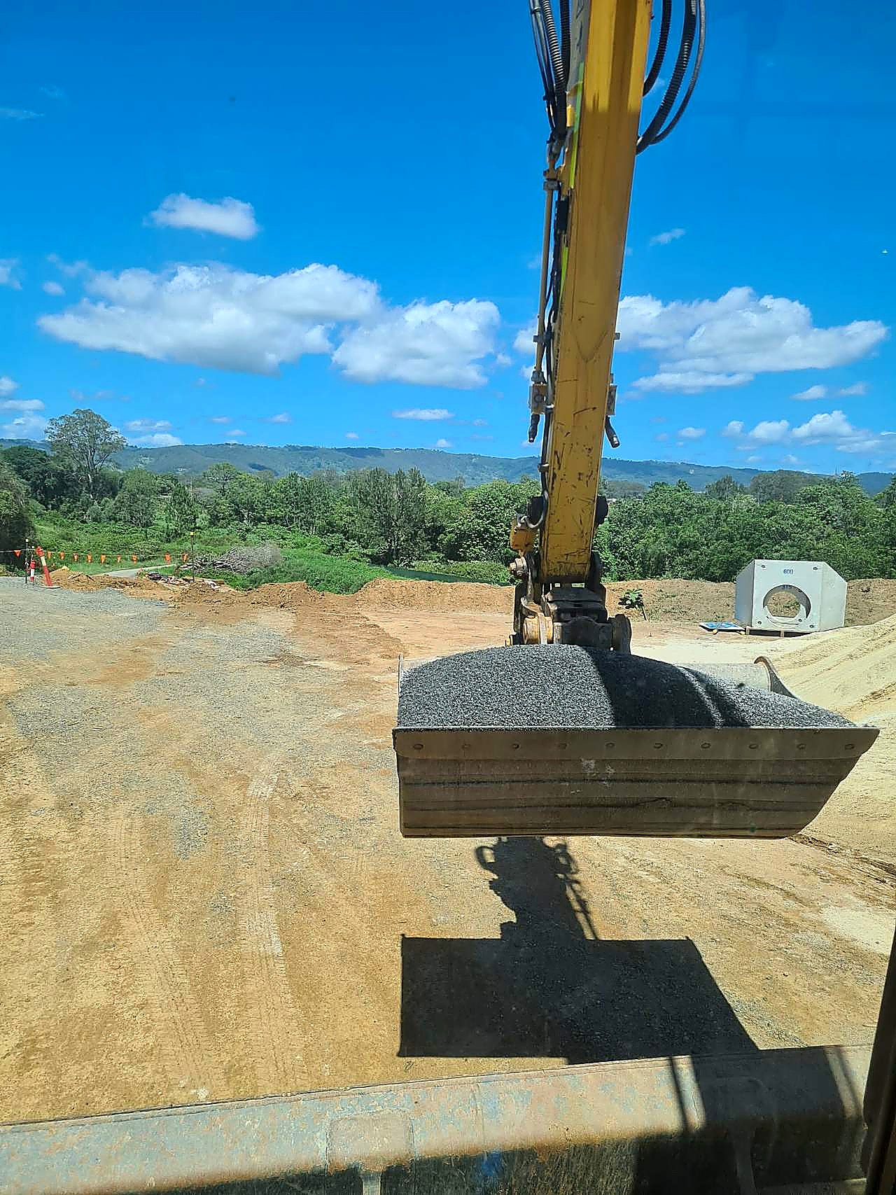 Yellow excavator carrying a bucket of crushed aggregate with a precast concrete pit on an SEQ civil site, photographed from the cab.